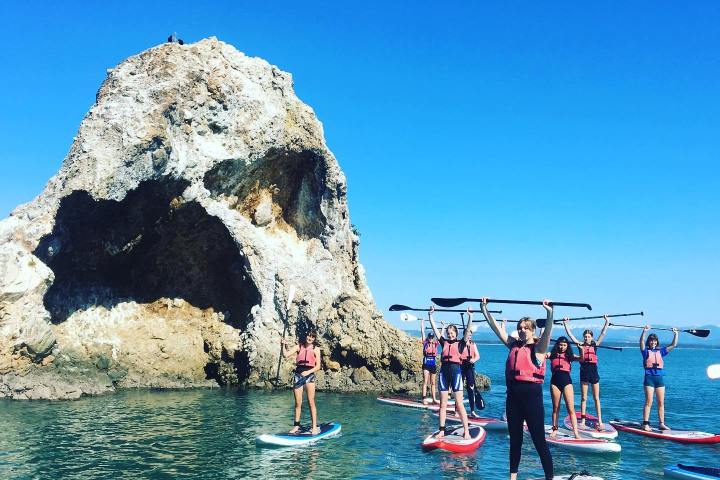 Group of people stand on paddleboards by a large rock in clear blue water, holding paddles above their heads.
