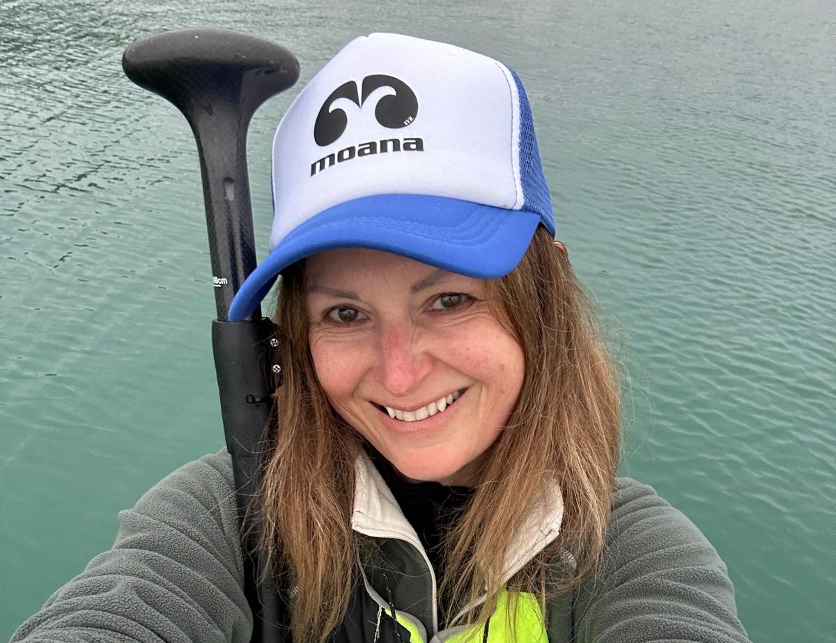 Woman on a paddleboard, smiling, wearing a cap and holding a paddle on a calm water body.